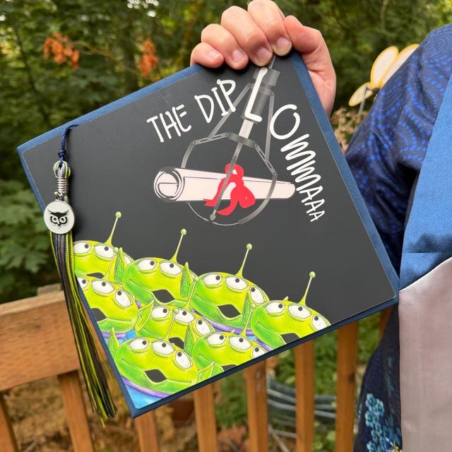 Graduate holding a Toy Story aliens 'The Diploma' grad cap topper