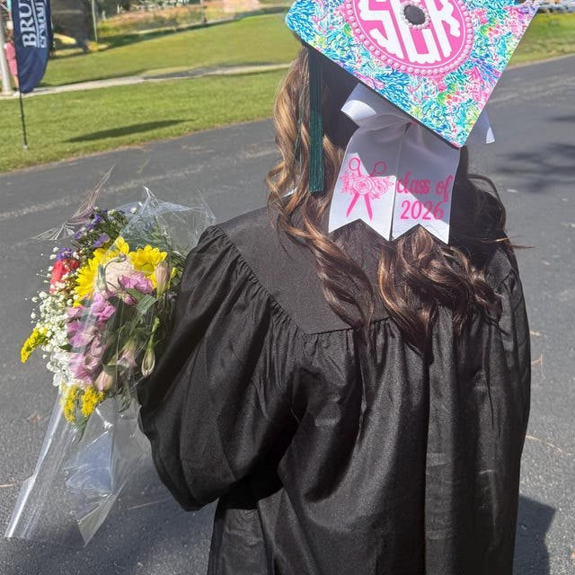 Graduate in a black gown holding flowers and wearing a colorful monogram grad cap topper