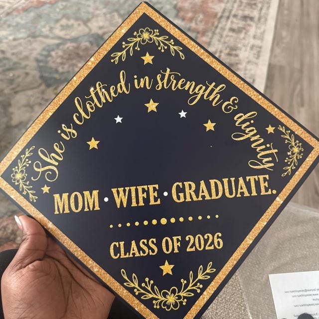 Graduate holding a 'Mom Wife Graduate Class of 2026' gold and black grad cap topper