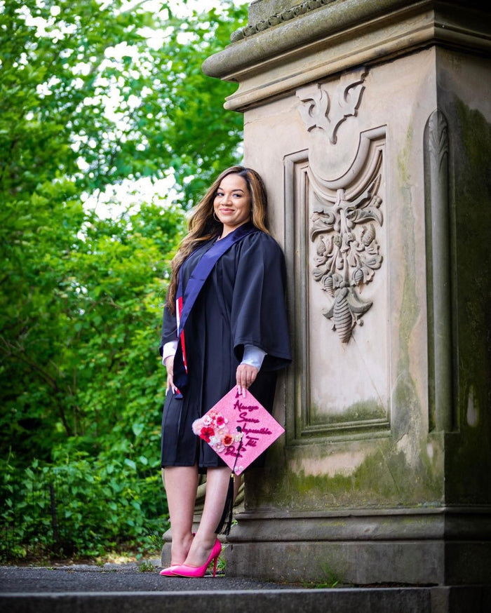 Graduate in a gown holding a pink Barbie grad cap topper outdoors