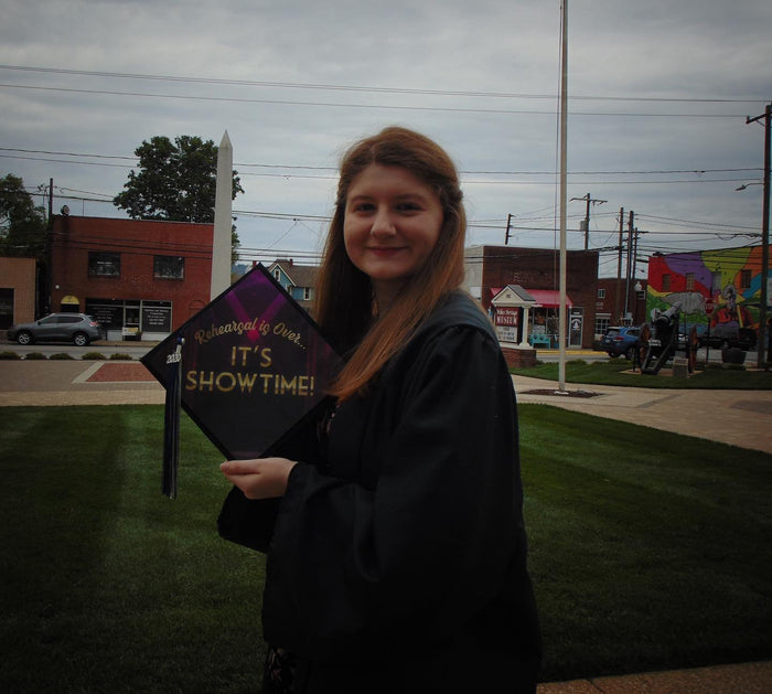 Graduate in a gown holding a Beetlejuice 'It's Showtime' grad cap topper outdoors