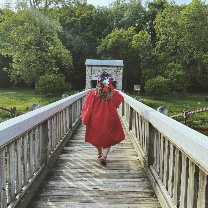 Graduate in a red gown wearing a floral grad cap topper walking across a wooden bridge