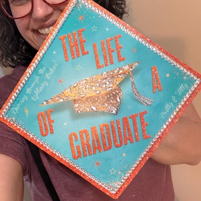 Graduate holding a 'The Life of a Graduate' turquoise and orange grad cap topper