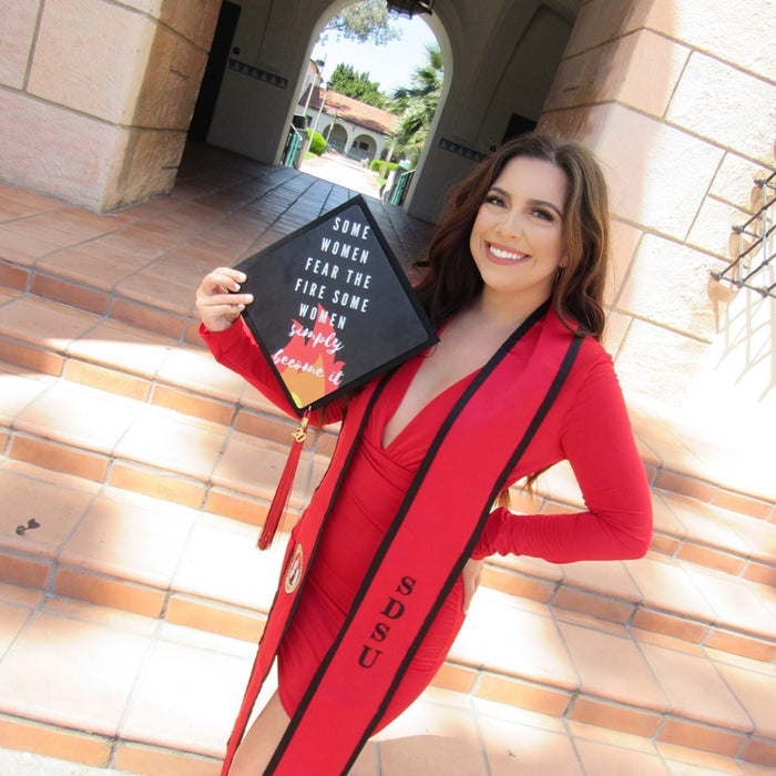 Graduate in a red gown holding a 'Some Women Fear the Fire' grad cap topper on campus steps