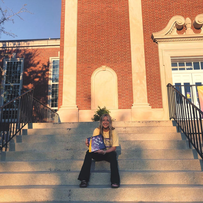 Graduate sitting on school steps holding a purple 'Off to My Next Dream' grad cap topper