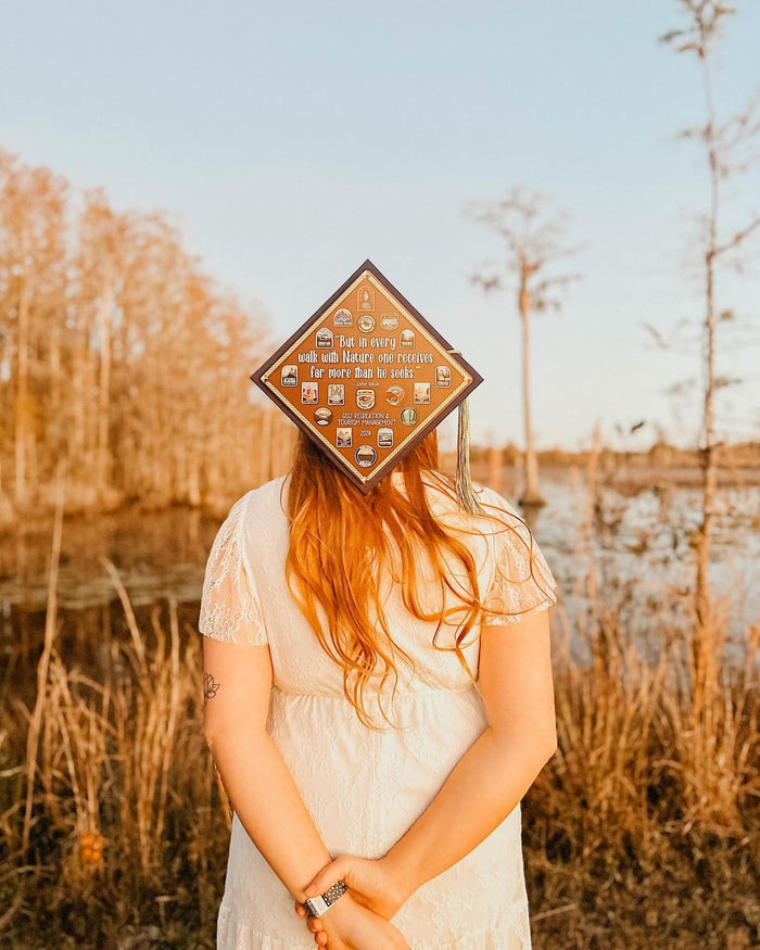 Graduate holding a national parks John Muir quote grad cap topper by a lake at sunset