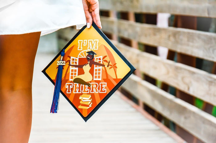 Graduate holding an 'I'm There' 2022 orange sunset grad cap topper on a bridge