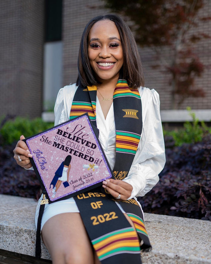 Graduate with a kente stole holding a 'She Believed She Mastered It' Class of 2022 grad cap topper