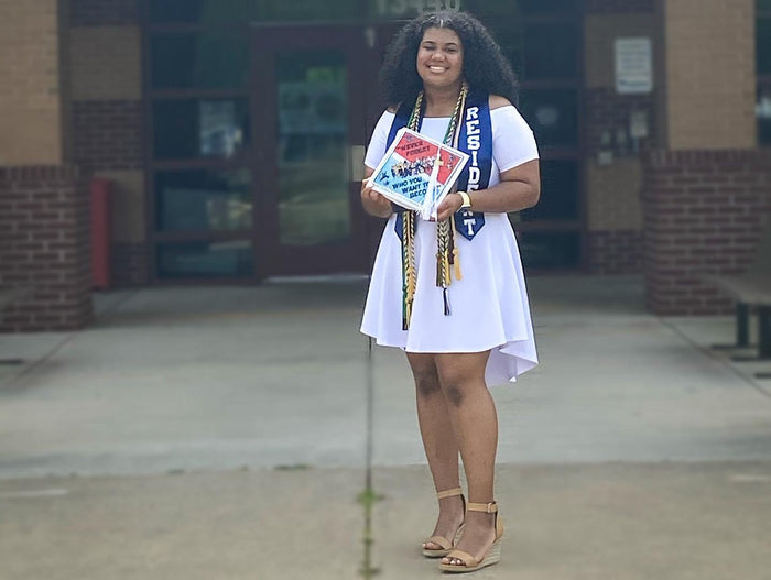 Graduate wearing a Resident sash holding a musical 'Never Forget Who You Want to Become' grad cap topper