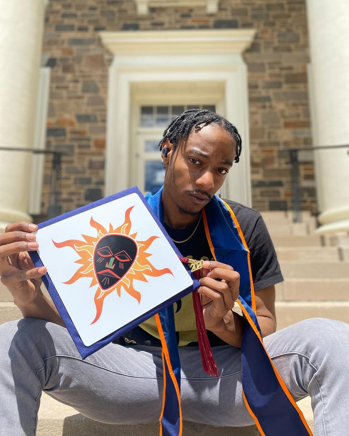 Graduate sitting on steps holding an African sun mask grad cap topper