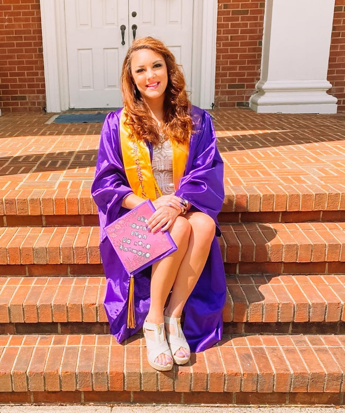 Graduate in a purple gown sitting on brick steps holding a pink glitter grad cap topper