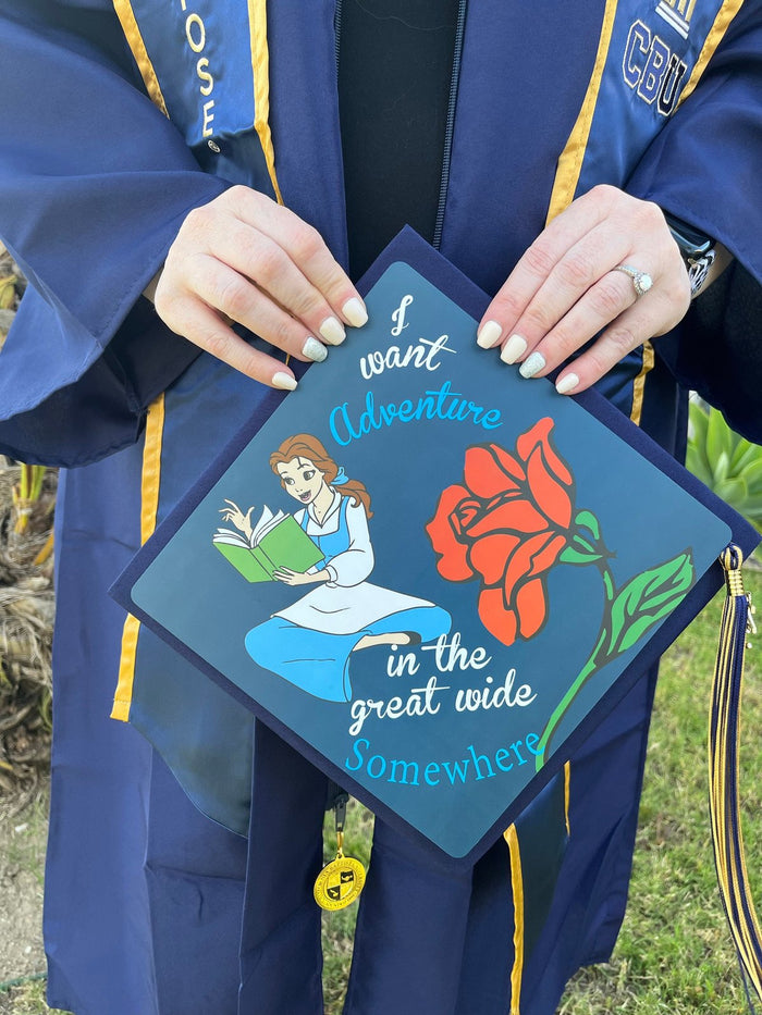 Graduate in blue gown holding Belle I Want Adventure topper