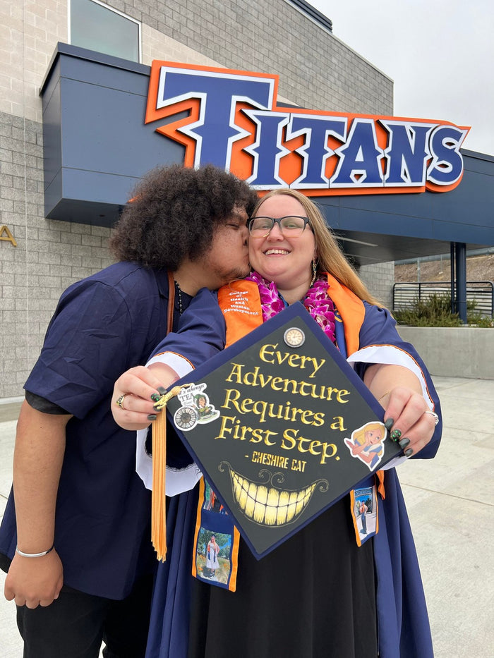 Graduate holding Cheshire Cat Every Adventure cap at Titans sign