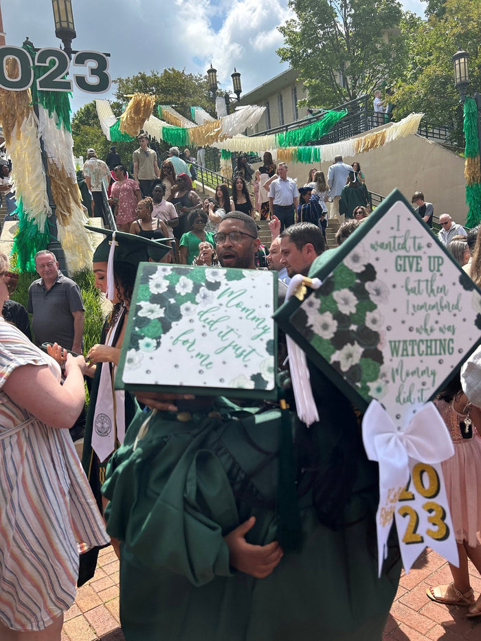 Two graduates holding matching floral Mommy Did It caps at ceremony
