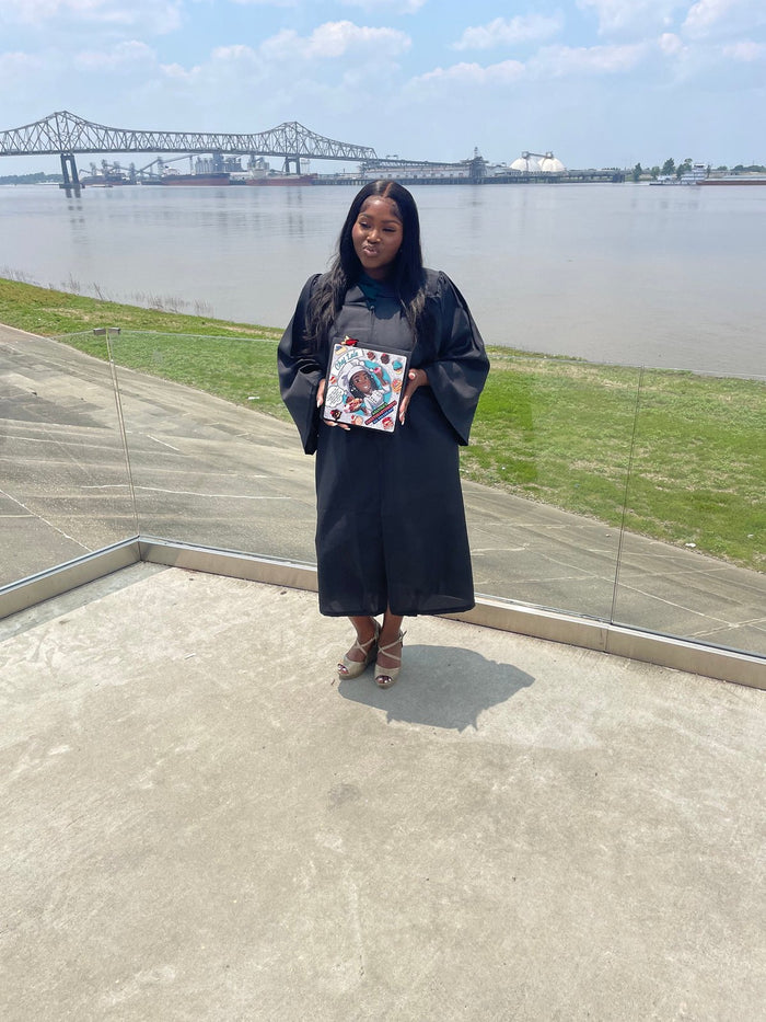 Graduate in gown holding Chef Lele cap by riverfront