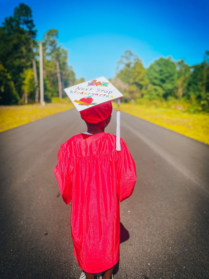 Child in red gown wearing Next Stop Kindergarten cap on road