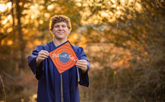 Graduate in blue gown holding Thats All Folks topper outdoors