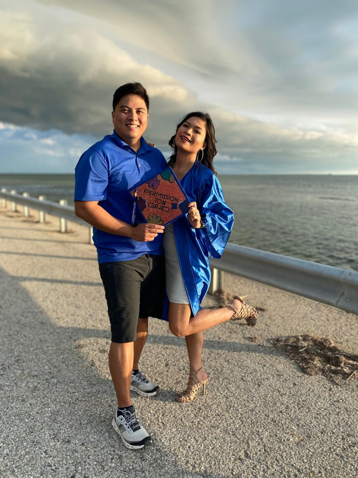 Graduate in blue gown holding Permission To Grad cap by the sea