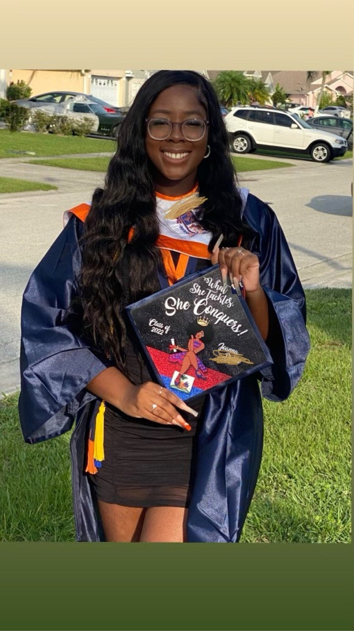Graduate in blue gown holding She Conquers cap