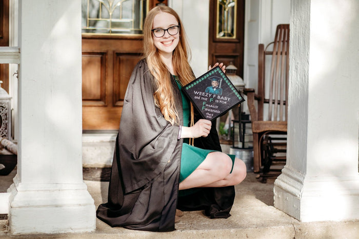 Graduate kneeling holding Weezy F Baby Finally Finished cap on porch