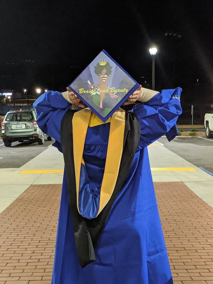 Graduate holding Brains And Beauty cap over face at night