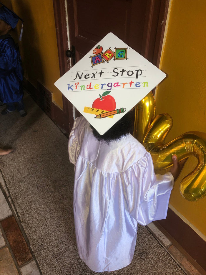 Child in white gown wearing Next Stop Kindergarten cap