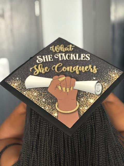 Woman holding What She Tackles She Conquers cap