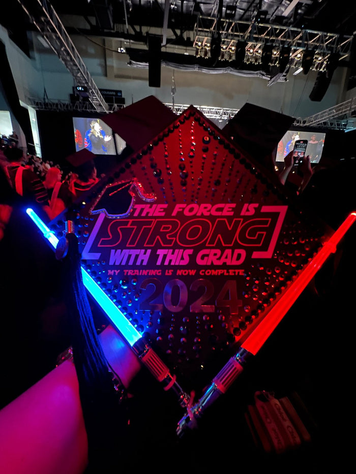 Graduate holding Force Is Strong 2024 cap with lightsabers at arena