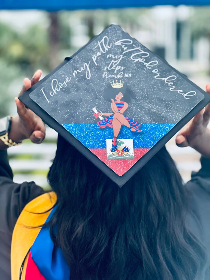 Graduate holding Haiti flag God Ordered My Steps cap